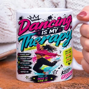 May include: A white ceramic mug featuring the text Dancing Is My Therapy in pink and blue. The design includes a graphic of a person dancing, musical notes, stars, and text lists including Clearer Mind, Stronger Body, Better Mood, More Focus, Happier Me.