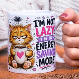 May include: A white ceramic mug featuring a graphic of a fluffy orange cat with a grumpy expression holding a small mug with a pink heart. The text reads I'M NOT LAZY I'M IN ENERGY SAVING MODE in black, pink, and purple lettering with small stars.