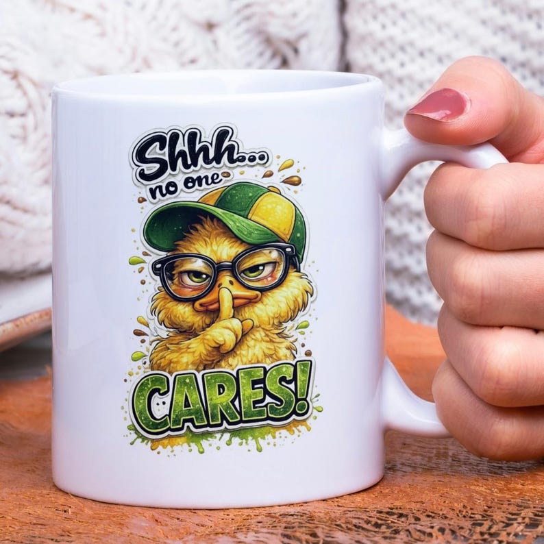 Op de afbeelding: Witte keramische mok met een cartoon eend met een bril en een baseballpet, met de tekst "Shhh... no one CARES!". De eend houdt een vinger voor zijn lippen. De mok heeft een wit handvat.