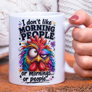 Op de afbeelding: Witte keramische mok met een kleurrijk, cartoonachtig hanenontwerp. De tekst op de mok luidt: "I don't like MORNING PEOPLE or mornings... or people..."