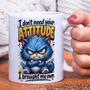 Op de afbeelding: Witte keramische mok met een illustratie van een chagrijnige blauwe kat en de tekst "I don't need your ATTITUDE I brought my own". De kat heeft gekruiste armen en gele ogen. De tekst is in zwart en geel.