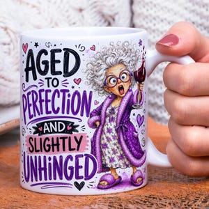 May include: A white ceramic mug featuring a cartoon illustration of an elderly person with gray hair, purple glasses, and a purple robe, holding a glass of red wine. The text reads AGED TO PERFECTION AND SLIGHTLY UNHINGED with small pink hearts.
