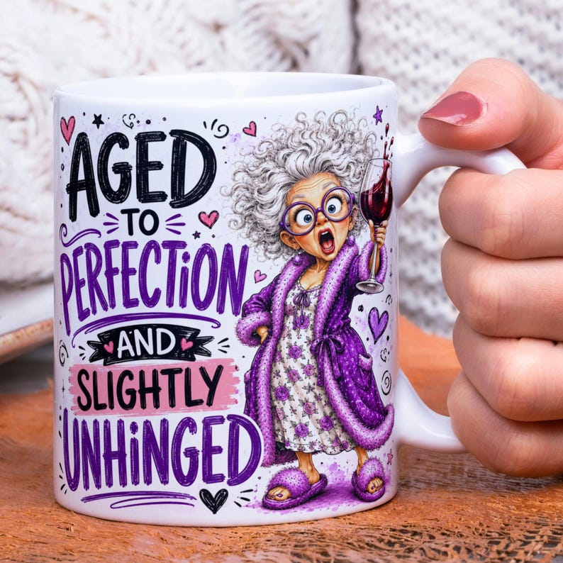 May include: A white ceramic mug featuring a cartoon illustration of an elderly person with gray hair, purple glasses, and a purple robe, holding a glass of red wine. The text reads AGED TO PERFECTION AND SLIGHTLY UNHINGED with small pink hearts.