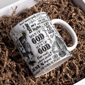 May include: White ceramic mug with a wraparound design featuring religious imagery and text. The mug includes illustrations of praying hands, a figure with a crown of thorns, and the words "MAN OF GOD" and "JESUS". The mug is in a white gift box with brown paper filler.