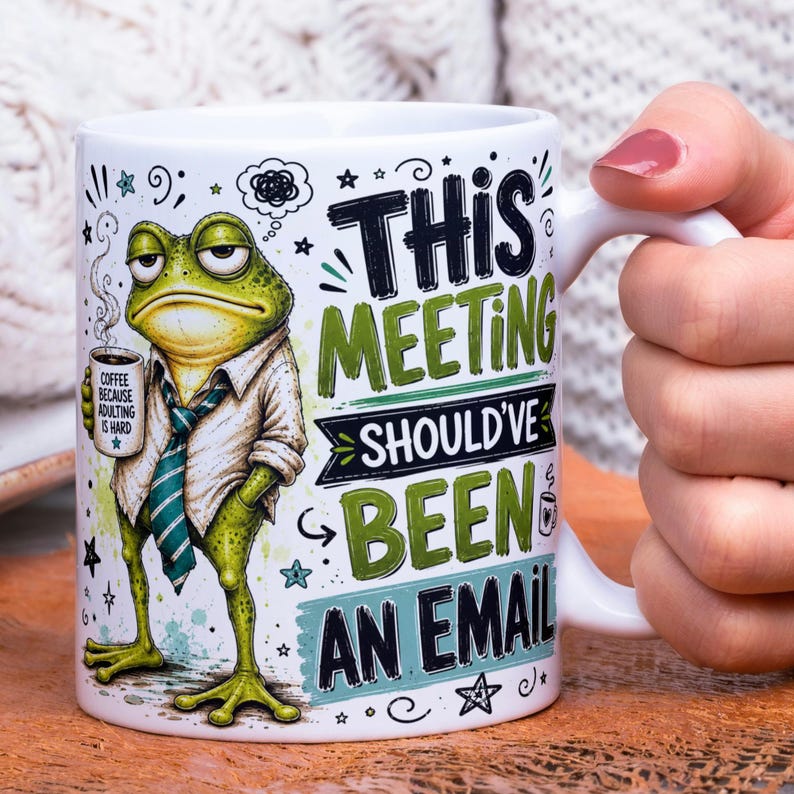 May include: A white ceramic mug featuring a green frog wearing a white shirt and a teal striped tie. The frog holds a small mug labeled COFFEE BECAUSE ADULTING IS HARD. The text THIS MEETING SHOULD'VE BEEN AN EMAIL is printed in black and green.