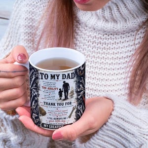 May include: A white ceramic mug with the text "TO MY DAD" and a graphic of a father and child walking. The mug has a black and white design with additional text. The mug is filled with a light brown liquid.