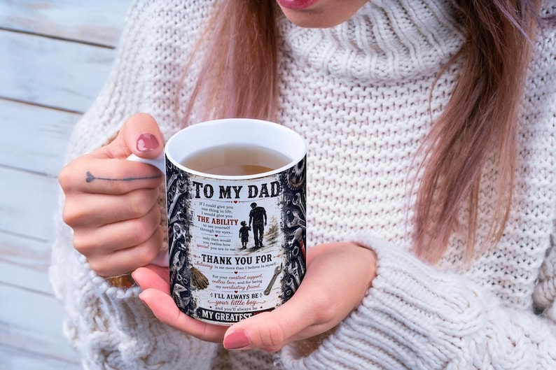 May include: A white ceramic mug with the text "TO MY DAD" and a graphic of a father and child walking. The mug has a black and white design with additional text. The mug is filled with a light brown liquid.