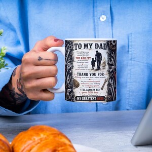May include: White ceramic mug with the text "TO MY DAD" and a graphic of a father and child. The mug also includes text that reads "THANK YOU FOR" and "MY GREATEST HERO". The mug is held by a person.