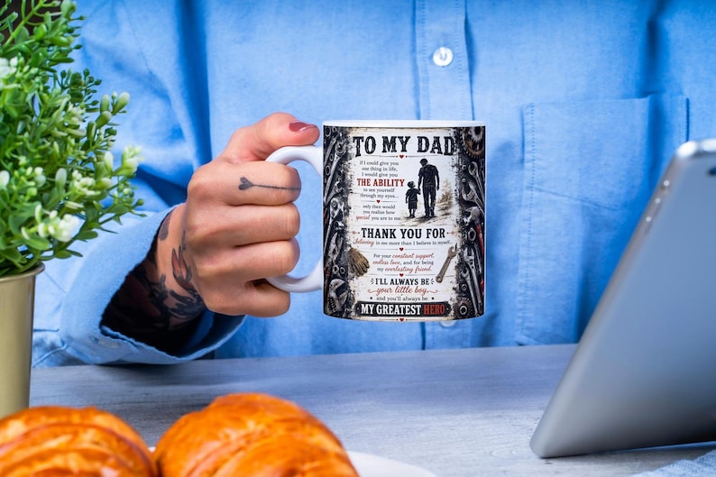 May include: White ceramic mug with the text "TO MY DAD" and a graphic of a father and child. The mug also includes text that reads "THANK YOU FOR" and "MY GREATEST HERO". The mug is held by a person.