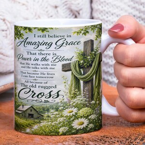 Op de afbeelding: Witte keramische mok met de tekst "I still believe in Amazing Grace" en een kruisontwerp, bloemen en een klein huis. De mok heeft een gebogen handvat en wordt door een persoon vastgehouden.