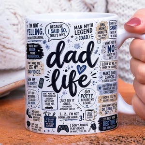 May include: White ceramic mug with the words "dad life" in a black script font. The mug is decorated with various phrases and illustrations related to fatherhood, including "I'm not yelling" and "Dad Fuel".