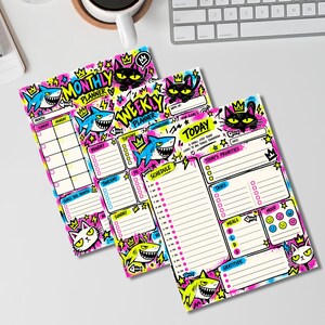 Puede incluir: Dos planificadores de papel con ilustraciones vibrantes estilo grafiti de tiburones y gatos con coronas. Los diseños incluyen textos como Monthly Planner, Weekly Planner y Today, con secciones para horarios, tareas, comidas y estado de ánimo.