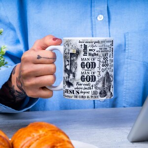 May include: White ceramic mug with religious text and imagery. The mug features the words "MAN OF GOD", "JESUS begins LORD", and other phrases. The design includes a lion, praying hands, and a cross. The mug is held by a person.