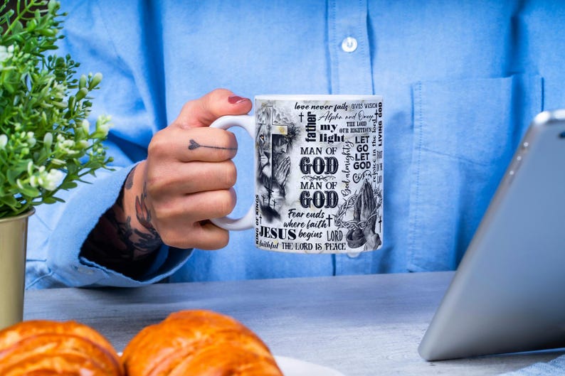May include: White ceramic mug with religious text and imagery. The mug features the words "MAN OF GOD", "JESUS begins LORD", and other phrases. The design includes a lion, praying hands, and a cross. The mug is held by a person.