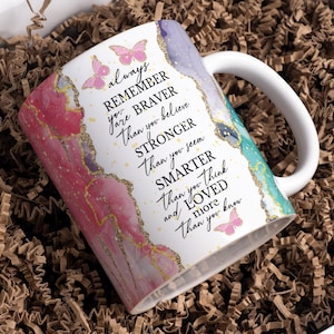 May include: White ceramic mug with a curved handle, featuring a colorful design with pink, purple, and teal watercolor-style accents. The mug has gold glitter accents and the text "always remember you're braver than you believe..." with butterfly illustrations.