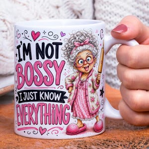 May include: A white ceramic mug featuring a cartoon illustration of an elderly person with gray curly hair, pink glasses, and a floral robe, holding a wooden rolling pin. The text reads: I'M NOT BOSSY I JUST KNOW EVERYTHING in black and pink.