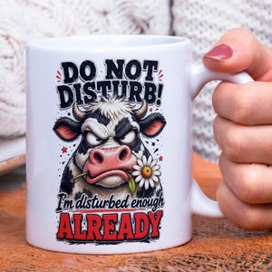 Op de afbeelding: Witte keramische mok met een zwart-wit illustratie van een koe. De koe heeft een boze uitdrukking en een madeliefje in haar mond. De tekst op de mok luidt "DO NOT DISTURB!" en "I'm disturbed enough ALREADY".