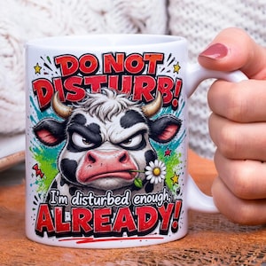 May include: White ceramic mug with a humorous design featuring an angry cow illustration. The mug has the text "DO NOT DISTURB!" and "I'm disturbed enough ALREADY!" in red and white. The cow is black and white with a flower in its mouth.
