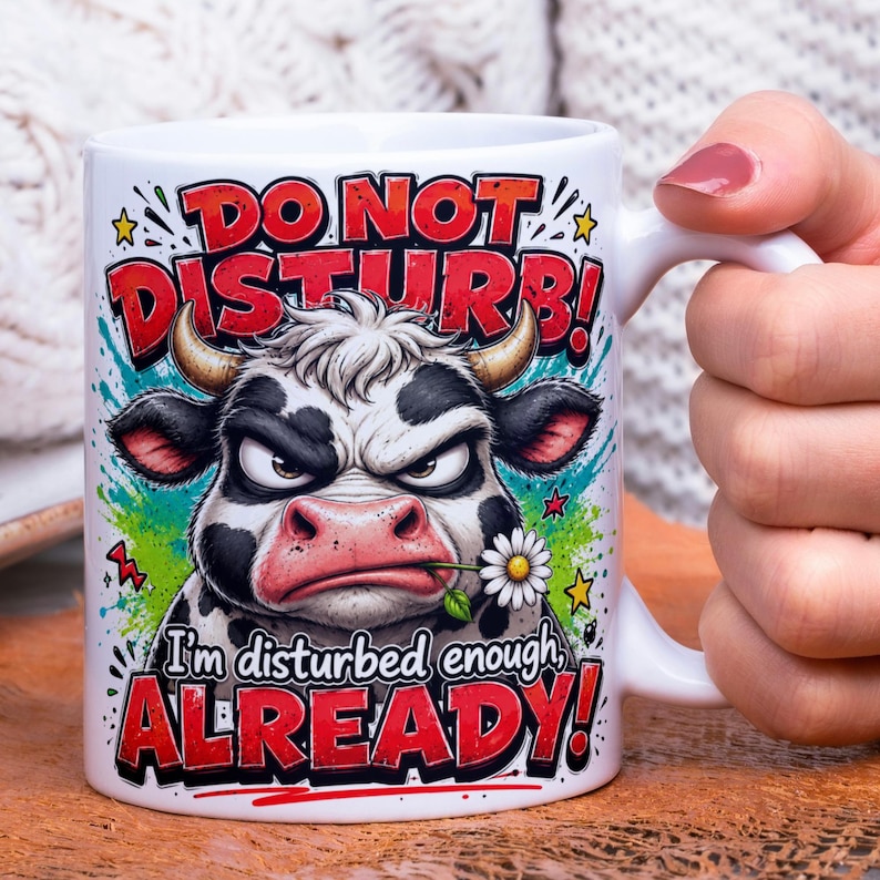 May include: White ceramic mug with a humorous design featuring an angry cow illustration. The mug has the text "DO NOT DISTURB!" and "I'm disturbed enough ALREADY!" in red and white. The cow is black and white with a flower in its mouth.
