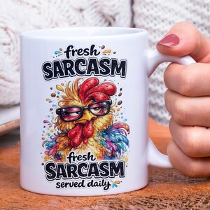 Op de afbeelding: Witte keramische mok met een humoristisch ontwerp. Het toont een cartoon haan met een bril, met de tekst "fresh SARCASM served daily". De haan is afgebeeld in levendige kleuren, waaronder geel, rood en blauw.