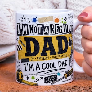 May include: White ceramic mug with the text "I'M NOT A REGULAR DAD I'M A COOL DAD" in black and yellow. The mug features illustrations of tools and a tape measure, with paint splatters and a crown.