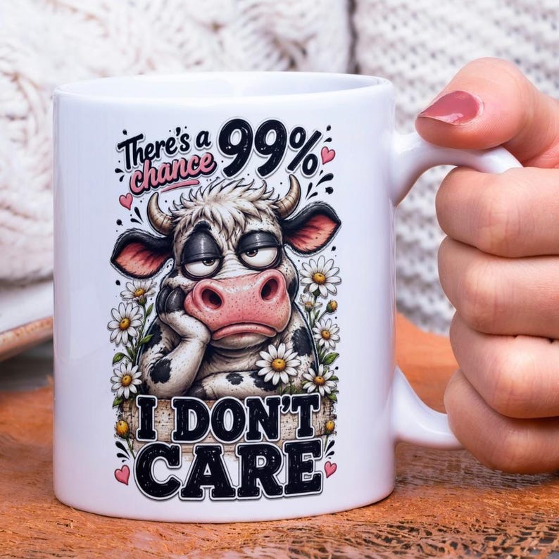 Op de afbeelding: Witte keramische mok met een cartoon koeienontwerp met de tekst "There's a 99% chance I DON'T CARE". De koe heeft een verveelde uitdrukking en is omringd door madeliefjes. De mok heeft een wit handvat.
