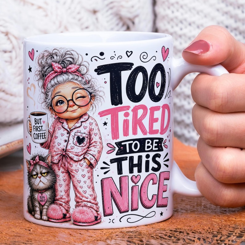 May include: A white ceramic mug featuring a cartoon illustration of a person in pink heart-patterned pajamas with a cat, wearing glasses and a headband. The text reads "TOO TIRED TO BE THIS NICE" in black and pink, with a small "BUT FIRST... COFFEE" mug.