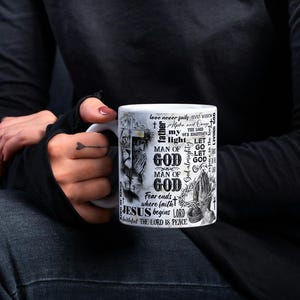 May include: White ceramic mug featuring religious text and imagery. The design includes the words "GOD", "JESUS", and "LORD", along with illustrations. The mug is held by a person wearing a black long-sleeve shirt and dark jeans.