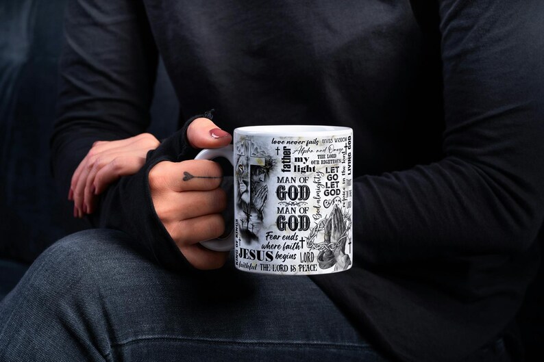 May include: White ceramic mug featuring religious text and imagery. The design includes the words "GOD", "JESUS", and "LORD", along with illustrations. The mug is held by a person wearing a black long-sleeve shirt and dark jeans.