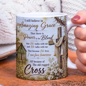 May include: White ceramic mug with a wraparound design featuring a wooden cross, floral accents, and text that reads "I still believe in Amazing Grace...Power in the Blood...The old rugged Cross." The mug has a white handle.
