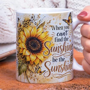 May include: White ceramic mug featuring a large yellow sunflower with brown and green accents, and butterflies. The mug has the text "When you can't find the sunshine, be the sunshine" in gold script.