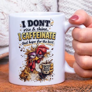 Op de afbeelding: Witte keramische koffiemok met een humoristisch ontwerp. De mok heeft een cartoonhaan met een slaperige uitdrukking die een koffiekopje vasthoudt. De tekst op de mok luidt: "I DONT rise & shine, I CAFFEINATE and hope for the best." Op het koffiekopje staat "COFFEE RIGHT MEOW."