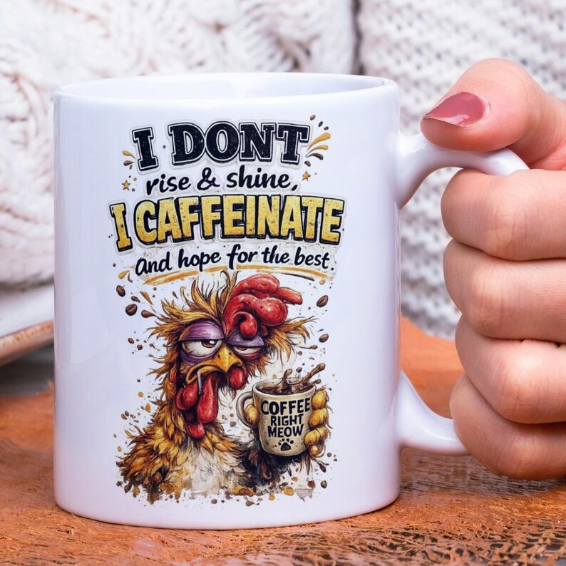 Op de afbeelding: Witte keramische koffiemok met een humoristisch ontwerp. De mok heeft een cartoonhaan met een slaperige uitdrukking die een koffiekopje vasthoudt. De tekst op de mok luidt: "I DONT rise & shine, I CAFFEINATE and hope for the best." Op het koffiekopje staat "COFFEE RIGHT MEOW."