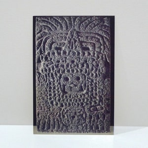 May include: A rectangular, gray stone carving with intricate relief details. The artwork features a stylized face, possibly a skull, and other geometric patterns. The stone has a rough, textured surface, suggesting an ancient artifact or historical piece.