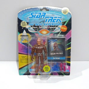 May include: A Star Trek: The Next Generation action figure of a Vogon alien, packaged on a colorful card. The figure is brown with a detailed design, and the packaging includes the text "NEW!" and "VORGON".