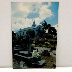 May include: A vintage postcard featuring a white church with a steeple, set against a backdrop of a blue sky with fluffy clouds. The church is surrounded by lush greenery and a cemetery with headstones and crosses.