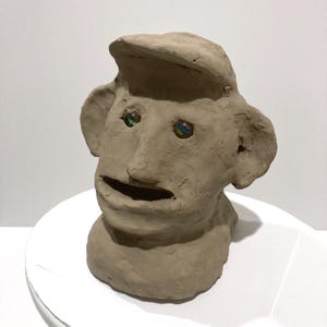 May include: A handmade clay sculpture of a human head with a hat. The sculpture has a rough texture and features glass marble eyes with colorful swirls. The mouth is open, and the ears are large. The sculpture is on a white surface.
