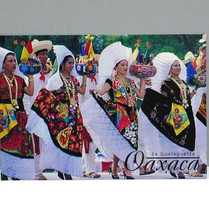 May include: A postcard featuring a group of people in traditional Mexican attire. The women wear colorful embroidered dresses, white lace skirts, and elaborate headdresses. The text "La Guelaguetza Oaxaca" is visible.