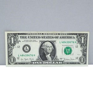 May include: A United States one-dollar bill. The bill features a portrait of George Washington and the text "The United States of America." The bill is green and black with the number "1" in each corner.