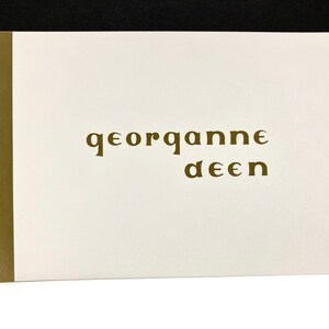 May include: A spiral-bound notebook with a white cover and a gold-colored spine. The cover features the name "georganne deen" in a stylized, gold-colored font. The notebook is open, displaying the blank pages.
