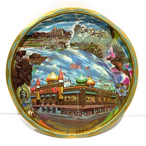 May include: A vintage round metal tray with a gold rim and a detailed illustration. The artwork depicts scenes of South Dakota, including Mount Rushmore, the Badlands, bison, and the Mitchell Corn Palace. Text includes "Mitchell, South Dakota".