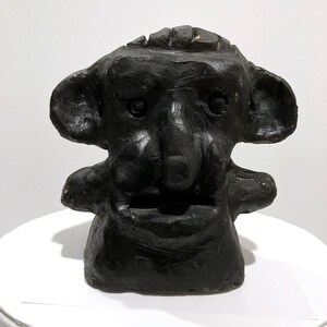 May include: A black, textured sculpture of a stylized face with large ears, a prominent nose, and a slightly open mouth. The sculpture is placed on a white surface, and the background is a plain white wall.