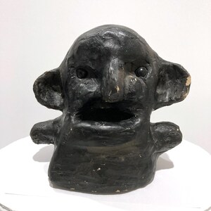 May include: A dark, textured sculpture of a face with large ears, a wide open mouth, and small arms. The sculpture is painted black and sits on a white surface. The face has a slightly distressed appearance.