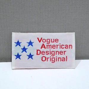May include: A rectangular white label with the text "Vogue American Designer Original" in red, and six blue stars. The label is likely for clothing or fashion items.