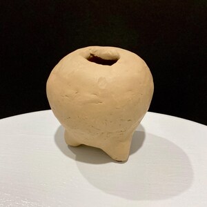May include: A small, beige ceramic vase with a rounded body and a single opening at the top. The vase has three short, rounded legs. The surface has a textured, handmade appearance.