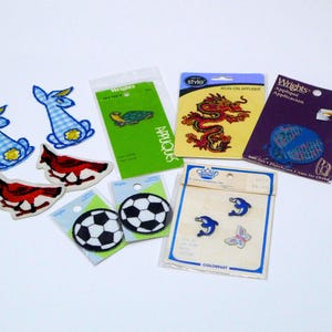 May include: A collection of iron-on appliques and sew-on patches. Designs include a red dragon, blue bunnies, soccer balls, cardinals, dolphins, a turtle, and a butterfly. The appliques and patches are in various colors and sizes.