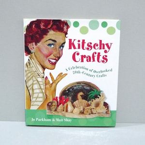 May include: A book titled "Kitschy Crafts" features a vintage illustration of a woman with red hair. The cover showcases a collection of seashells and coral. The subtitle reads "A Celebration of Overlooked 20th-Century Crafts."
