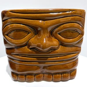 May include: A brown ceramic tiki mug with a square shape and a carved face design. The mug features a glossy finish and a detailed depiction of a tiki god with closed eyes, a wide nose, and a toothy grin. The mug is a decorative item.