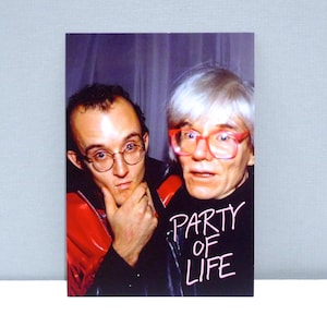 May include: A photograph featuring two men. One man wears glasses and a red jacket. The other man has white hair and red glasses, and a black shirt with the words "PARTY OF LIFE" in white. The background is a muted purple.