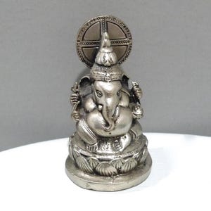 May include: A silver-colored statue of the Hindu deity Ganesha, seated on a lotus flower base. The statue features intricate details, including a large elephant head, multiple arms, and a decorative halo behind the head. The statue is approximately 4 inches tall.